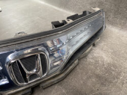 Close-up of a car's front end showing a Honda emblem and a detached headlight assembly on the workshop floor.