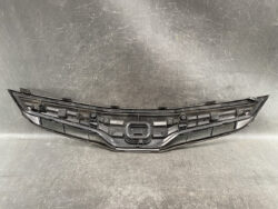 Black plastic front bumper grille with mounting clips and central badge area, shown on a workshop bench.