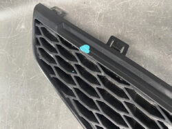 Black plastic car grille with a diamond mesh pattern resting on a dirty surface, with a small turquoise sticker on it.