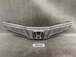 Front Honda car grille with honeycomb mesh and central logo, shown as a spare part on a workshop surface