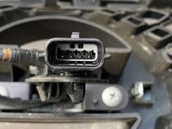 Six-pin automotive electrical connector with a rectangular black housing and visible metal pins inside, mounted to a harness edge.