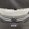 Front view of a Nissan car grille with chrome slats and circular Nissan badge, plus a light-colored hood panel resting above it. NISSAN NOTE E12 Genuine Radiator Grille 62310 5WC0A » JDM-PARTS NZ