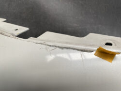 Close-up of a scratched, dented car panel along a seam, with a small piece of yellow tape near the edge on light gray paint.