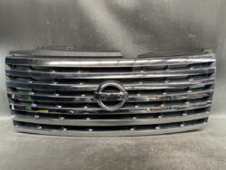 Front grille of a Nissan vehicle with chrome horizontal slats and circular Nissan emblem in the center, against a dark background.