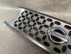 Close-up of Nissan emblem on a black honeycomb car grille, showing a metallic logo and hexagonal openings.