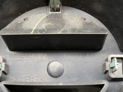 Close-up of a Nissan plastic car part with part number 62310 JG00A, showing a circular access plug and ABS markings on a dusty surface.