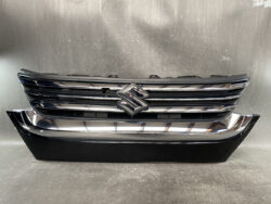 Front view of a Suzuki car grille with chrome horizontal bars and logo centered, mounted on a black bumper cabinet.