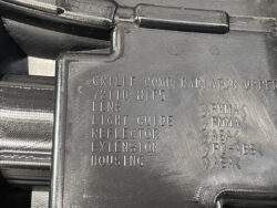 Close-up of a black plastic automotive housing with embossed part labels: Grille Comp, Radiator Upper, Lens, Light Guide, Reflector, Extension, Housing; material codes on the right (PMMA, ASA, PC-ABS).