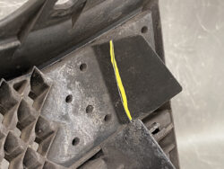 Close-up of a dirty black plastic panel with a yellow scratch running along the edge, suggesting damage to a vehicle trim piece.