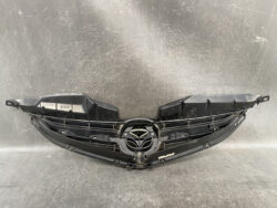 Black Mazda front grille assembly with central logo, photographed on a scratched gray surface