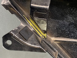 Close-up of a black metal bracket with a yellow seam or gasket along a joint, showing a notch and a bolt hole area.