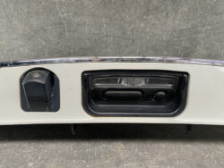 Close-up of a car's rear bumper showing the trunk latch handle and a small circular lock on a gray panel.